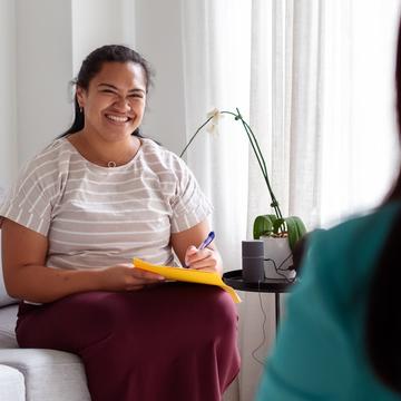 Smiling woman talking to another person while sitting on a couch and writing in a notebook
